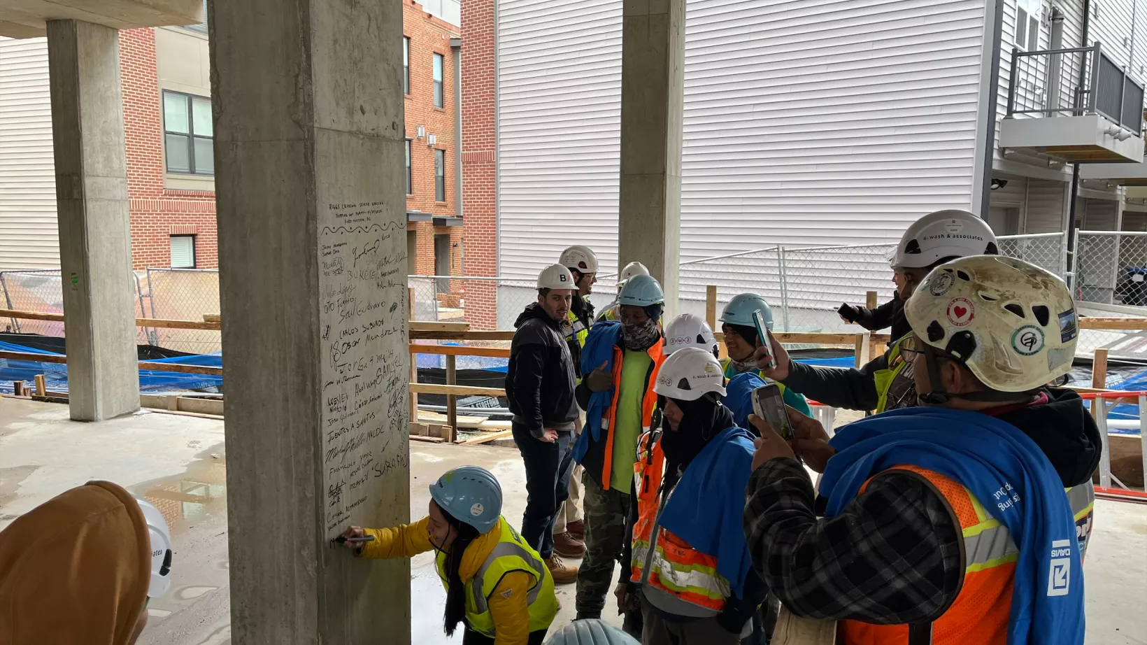 A group of people sign a pillar within a construction site, marking the construction team reaching the top of the structure.