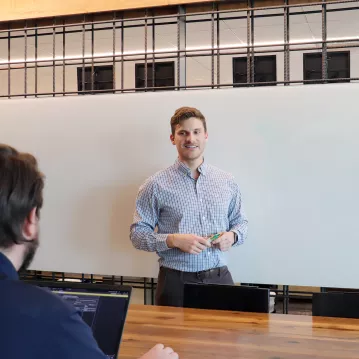 A man in professional wear stands in front of a whiteboard. Another man with a computer sits at a desk in front of him.