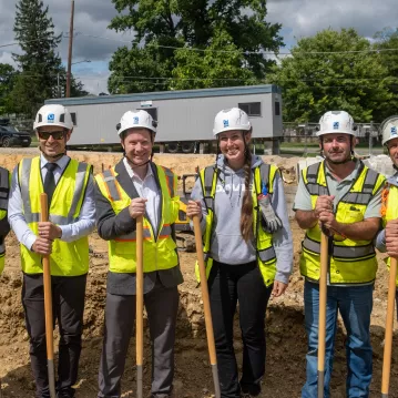 People in construction gear pose and hold shovels at a groundbreaking ceremony.