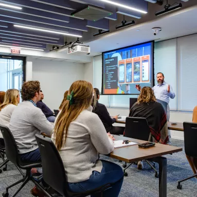 Two rows of people sit in a classroom as one man stands at the front and delivers a lecture.