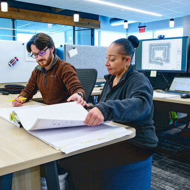 A man and woman sit at a desk in discussion, flipping through pages of a document.