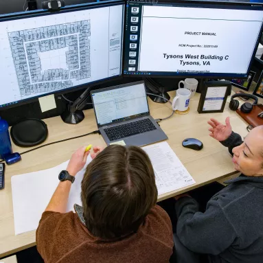 A man and woman sit at a desk in discussion, with documents displayed on the monitors in front of them.