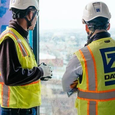 Two men wearing construction gear stand in an ongoing project and look out of a window.