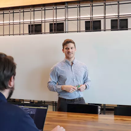 A man in professional wear stands in front of a whiteboard. Another man with a computer sits at a desk in front of him.