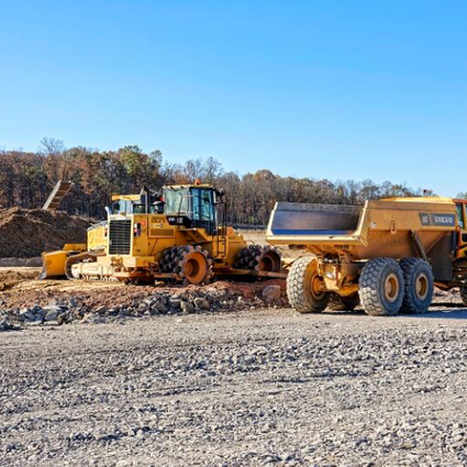 Two construction vehicles ride through a construction site, over gravel and dirt.