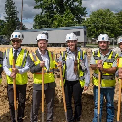 People in construction gear pose and hold shovels at a groundbreaking ceremony.