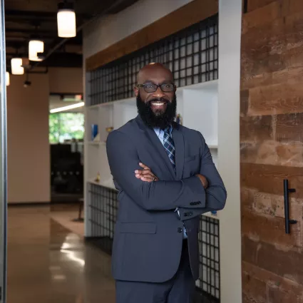 A man in a suit stands with arms crossed in office hallway