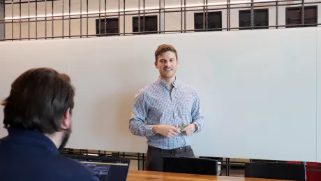 A man in professional wear stands in front of a whiteboard. Another man with a computer sits at a desk in front of him.