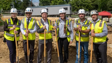 People in construction gear pose and hold shovels at a groundbreaking ceremony.