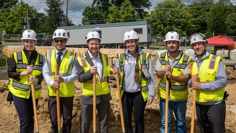 People in construction gear pose and hold shovels at a groundbreaking ceremony.