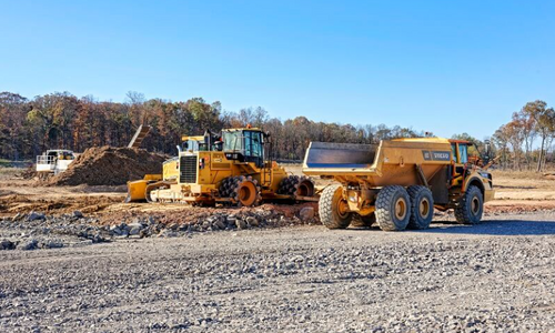 Two construction vehicles ride through a construction site, over gravel and dirt.