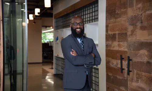 A man in a suit stands with arms crossed in office hallway