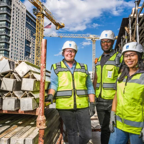 A group of people (two women and one man) stand together in construction gear next to equipment.