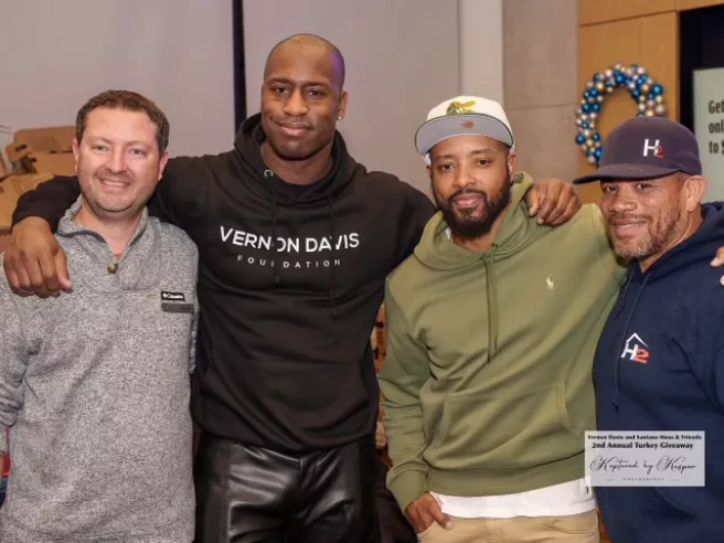 Four men (two of them football players) stand and pose for a picture at a Thanksgiving turkey giveaway event.