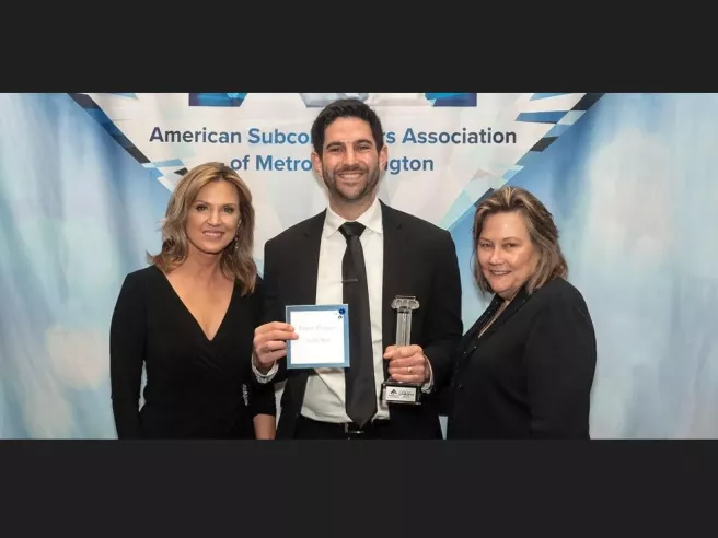 A group of people (two women and one man) stand together. The man in the middle holds an award. 